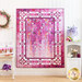 A full pink, purple and white ombre quilt with an elaborately quilted border hanging on a white paneled wall with white shelves holding home decor items and purple flowers on either side.