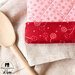 Close up of the red trim at the bottom of the oven mitt resting on a pile of natural colored towels and a wooden spoon angled to the left