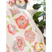 Close up of a cream quilt with abstract roses made of layered shades of pink fabric draped over furniture with home decor on a white shelf and a green houseplant in the background.