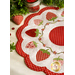 A white countertop with foliage and a houseplant in the background with a white and red strawberry themed table topper with a strawberry shaped pin cushion and a jar of red buttons in the background