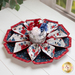 Flower shaped table topper with central opening with a small jar and patriotic decor in the background. Topper is made of crisp white, red and navy blue fabrics with multicolor florals.