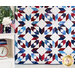 photo of a geometric patriotic-themed quilt hung vertically against a wall with a white flower, small shelf with patriotic decor and a green houseplant on one side..