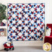 photo of a geometric patriotic-themed quilt hung vertically against a wall with a white flower, small shelf, red wagon decor and a green houseplant on one side and a white chair draped with a red blanket and holding a vase filled with flowers on the right.