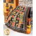 Large autumn themed quilt draped over furniture with a pumpkin and small stool with flowers on the floor in front of a small wooden shelf with a lantern and decor on it in front of wooden double doors.