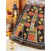 Large autumn themed quilt draped over furniture in front of a stone wall and small wooden shelf with a lantern and decor on it in front of wooden doors.