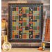 A large, autumn themed quilt in green, orange, black, and yellow with pumpkin and oak leaf applique details hanging on a wood paneled wall with a wooden ladder and orange fabric draped to the right and a small wooden crate with autumn decor on the left
