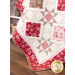 Close up photo of one corner of a red, pink, and cream quilt made with traditional style quilt star blocks and diamond blocks draped against a white paneled wall with a white shelf in the background, laden with flowers and a basket of coordinating yarn..