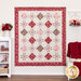 Photo of a red, pink, and cream quilt made with traditional style quilt star blocks and diamond blocks hanging flat against a white paneled wall with white furniture and red and pink decor like a blanket, books, roses, and yarn.