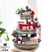 A tiered metal stand with patriotic mini pillows on every level with some plant decor in the background and a bright window and white paneled wall behind.