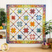Colorful quilt with diamonds all over with traditional style sawtooth stars made with floral fabrics from the Wild Blossoms collection hanging on a white paneled wall with houseplants on either side and a small white shelf with bee and floral decor
