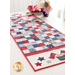 Photo of a patriotic red white and blue table runner in a patchwork pattern on a white table with a stack of plates and matching napkins, a patriotic centerpiece with a small American flag sits to the side in front of a bright window in the background
