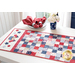 Photo of a patriotic red white and blue table runner in a patchwork pattern on a white table with a stack of plates and matching napkins, a patriotic centerpiece with a small American flag sits to the side in front of a bright window in a blue wall in the background