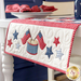 table runner hanging off the edge of a white table with a stack of plates and matching napkins in the background. close up shows quilting detail on one end with applique flags, stars, and a drum