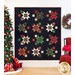Photo of a black quilt with red, green, and cream star bursts hanging on a white paneled wall with a decorated Christmas tree to the left with wrapped gifts and a chair with a red blanket and poinsettias atop it with a wreath hanging on the wall.