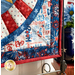 close up of patriotic quilt square made with red, white, blue, and yellow fabrics with stars and textures in a radiating pattern on a countertop with patriotic decor in the background