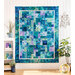 A blue, green, and purple quilt hanging on a white paneled wall with a houseplant and small white shelf with matching decor on either side.