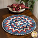 Image of a red, white, and blue circular table topper with a miniature quilt block center piece. The table topper is set on a brown wood table with spotted cloth napkins atop a stack of plates to the side