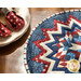 Image of a red, white, and blue circular table topper with a miniature quilt block center piece. The table topper is set on a brown wood table with spotted cloth napkins atop a stack of plates to the side