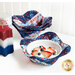A red, white, and blue bowl cozy made with patriotic fabrics holding a white bowl filled with fruit and whipped cream on a white table with a stack of matching bowl cozies in the background with a patriotic candle on the left.