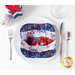 A red, white, and blue bowl cozy made with patriotic fabrics sitting on a plate on a white table top with patriotic candles, silverware, and a glass