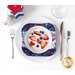 A red, white, and blue bowl cozy made with patriotic fabrics holding a white bowl with fruit and whipped cream inside sitting on a plate on a white table top with patriotic candles, silverware, and a glass