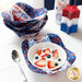 A red, white, and blue bowl cozy made with patriotic fabrics holding a white bowl filled with fruit and whipped cream on a white table with a stack of matching bowl cozies in the background with two patriotic candles on the right.