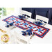 Patriotic themed red white and blue table runner on a white table with four place settings with matching cloth napkins all around with four white chairs and a bright window and blue wall in the background