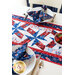 Close up of patriotic table runner showing a pinwheel quilt block pattern with a matching place setting on either side with a bowl of strawberries at the center of the table.