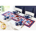 Patriotic themed red white and blue table runner on a white table with four place settings with matching cloth napkins all around and a white bowl of strawberries at the center with four white chairs and a bright window and blue wall in the background