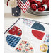 Close up of table runner detail showing stitching and fabric motifs with an American flag centerpiece and stack of plates with matching cloth napkins in the background