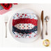 A red, white, and blue bowl cozy made with patriotic fabrics sitting on a plate on a white table top with patriotic decor, silverware, and a glass