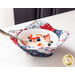 A red, white, and blue bowl cozy made with patriotic fabrics holding a white bowl filled with fruit and whipped cream on a white table with a silver spoon in the bowl.