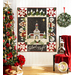 A black, white, and red quilt with a church in the center and village motifs around the border hanging on a white paneled wall with a chair with a red draped blanket on it, and a decorated christmas tree with wrapped gifts on the left