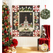 A black, white, and red quilt with a church in the center and village motifs around the border hanging on a white paneled wall and staged with a chair with a red draped blanket on it and a decorated christmas tree with wrapped gifts on the left.