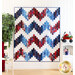 A red, white and blue quilt with a large zig zag pattern hanging on a white paneled wall with a small white chair and shelf with patriotic decor and a large houseplant on one side.