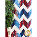 A red, white and blue quilt with a large zig zag pattern hanging on a wall with a large houseplant on one side.