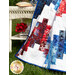 Gathered red white and blue quilt next to a white wicker table with red flowers and a book and green grass in the background