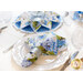 Angled photo of blue and white floral cloth napkin in a silver napkin ring on a white decorative plate with a matching table topper in the background made with fabrics from the same collection