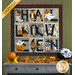Photo of a small Halloween quilt hanging on a green paneled wall with fall/Halloween decor and a gray table with drawers. Each block of the quilt contains a letter of the word 