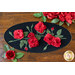 A black oval table topper with 3-dimensional roses and green leaves with gold beads around the edge on a brown wooden table with red roses on the top and bottom