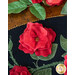 A black oval table topper with 3-dimensional roses and green leaves with gold beads around the edge on a brown wooden table with red roses on the top