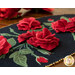 A black oval table topper with 3-dimensional roses and green leaves with gold beads around the edge on a brown wooden table with red roses in the background