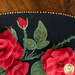 A black oval table topper with 3-dimensional roses and green leaves with gold beads around the edge on a brown wooden table