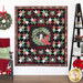 A finished Christmas Eve Quilt hanging on a white paneled wall with a ladder shelf on the right featuring homey decor, and a chair with a red blanket and green pillow on the other with a wreath hanging on the wall.