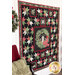 A finished Christmas Eve Quilt hanging on a white paneled wall with a chair and a red blanket with green pillow on top with a wreath hanging on the wall.