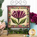Photo of a completed quilt block featuring a stylized flower with a purple butterfly accent made with metallic fabrics and thread hanging from a craft holder with swirls standing on a countertop against a white paneled wall with floral decorations all around