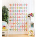Photo of a multicolored quilt with alternating white diamonds all over, hanging on a white paneled wall with a white wicker chair, houseplant, and floral decor on one side and a small white shelf with flowers in vases on the other