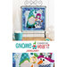 Photo of a finished wall hanging with a gnome building a snowman with snowflake motifs hanging on a metal craft hanger with a mug of cocoa and snowflake decor on either side and in front of a white paneled wall with the words 