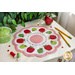 Scalloped table topper on a white counter featuring a ring of red and green apples on a white background with the alphabet and numbers in an embroidered ring in the middle. Pencils, spools of thread, a pot, buttons, and greenery are on the countertop 