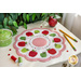 Scalloped table topper on a white counter featuring a ring of red and green apples on a white background with the alphabet and numbers in an embroidered ring in the middle. Pencils, spools of thread, a pot, buttons, and greenery are on the countertop.